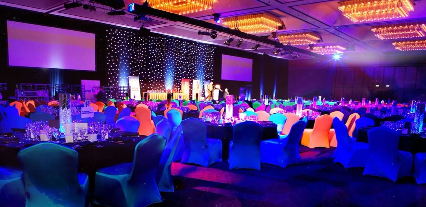 A gala ballroom with a star-cloth drape, colored uplighting on the walls, covered chairs and projection screens
