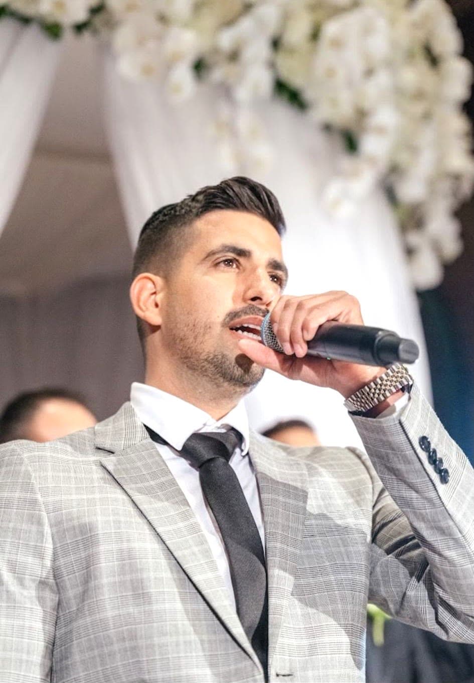 Ofek Ben Harush in a light gray suit, singing into a microphone in front of a white floral wedding canopy