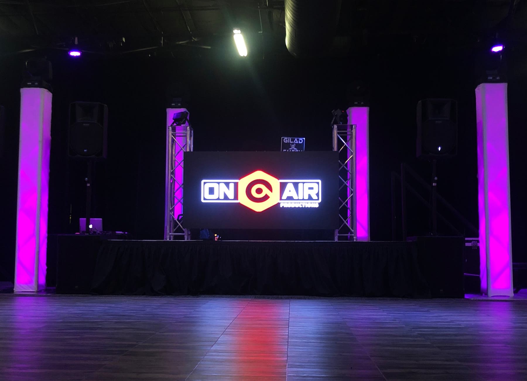 A DJ booth with a glowing logo screen in a darkened room, flanked by speakers and truss towers, with columns uplit in purple